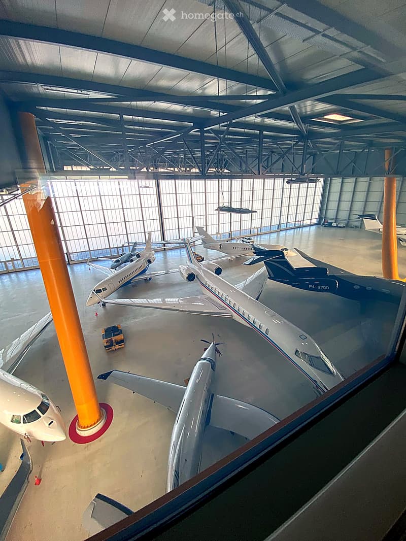 Hangar with multiple private jets parked inside, large windows, metal framework, and industrial lighting.