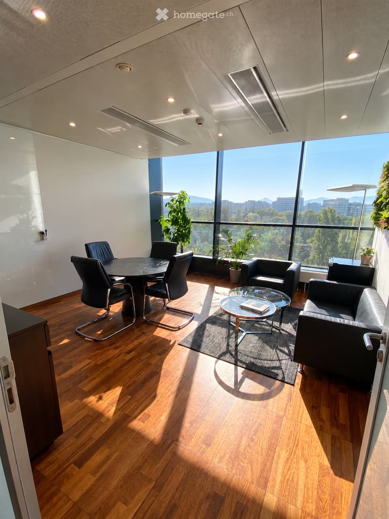 Modern office or meeting room with wooden flooring, large windows offering a city view, a round table with chairs, a sofa, a coffee table, and plants.