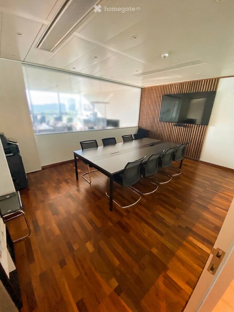 Modern conference room with a long rectangular table, multiple chairs, and a large screen on the wall. The room features wooden flooring and large windows with a city view.