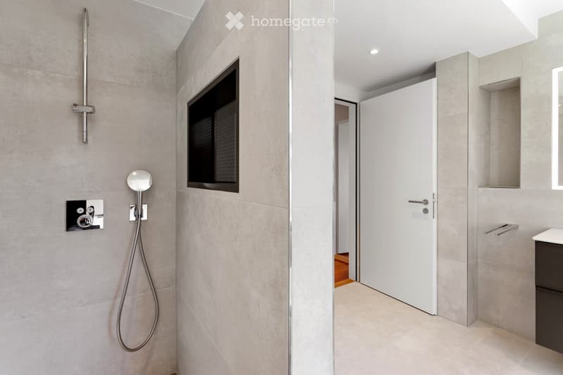 Modern bathroom with grey tiled walls, a sleek showerhead, a built-in mirror, and a dark cabinet.