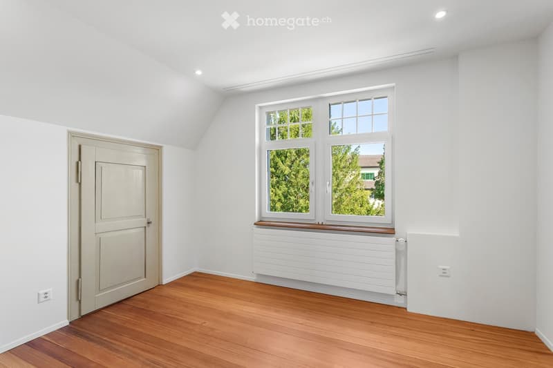 Empty room with white walls, wooden floor, large windows, radiator, and a door.