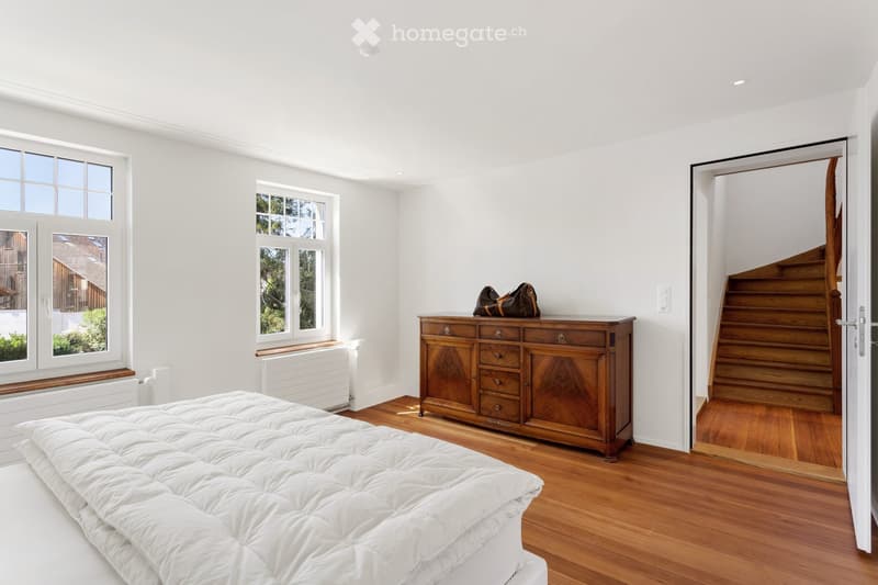 Bright bedroom with wooden flooring, large windows, a neatly made bed, a wooden dresser with drawers, and a handbag on top.