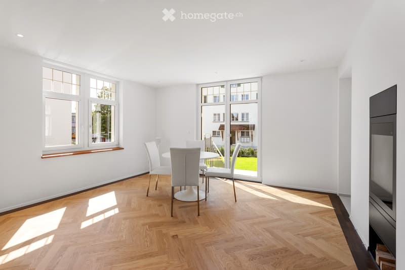 Bright, minimalist dining area with white walls, wooden herringbone flooring, and large windows. A white dining table with four matching chairs is positioned near the window, allowing natural light to fill the space.