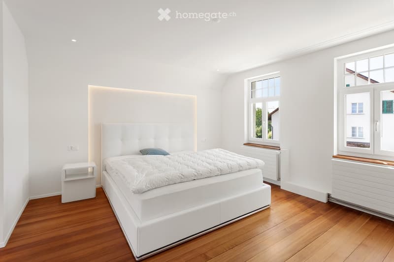 Modern bedroom with white walls, wooden floor, large bed with white headboard, nightstand, and windows with wooden sills.