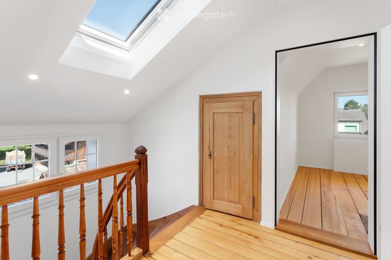 Bright, airy hallway with wooden flooring, white walls, and a skylight at the top. Features a wooden staircase with a handrail and a closed wooden door leading to another room.