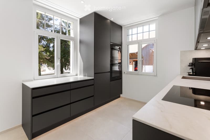 Modern kitchen with black cabinets, white countertops, and large windows offering a view of greenery outside.