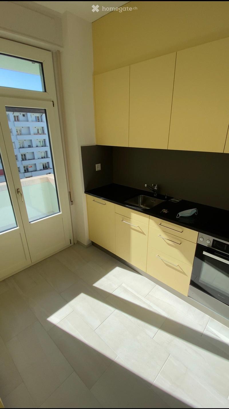 Modern kitchen with light yellow cabinets, black countertops, and a stainless steel oven. The space is well-lit with natural light coming through a large window and door leading to a balcony.