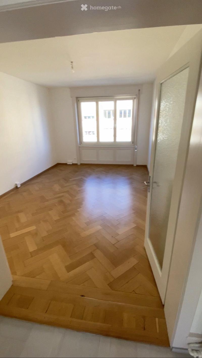 Empty room with wooden herringbone flooring, white walls, and a large window with white frame. No furniture or equipment present.