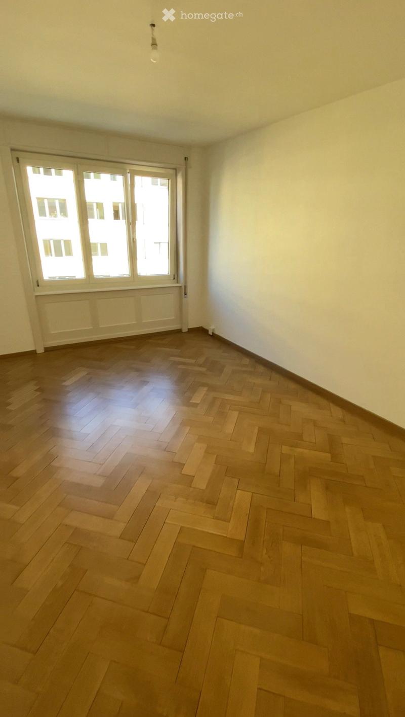 Empty room with wooden herringbone flooring, white walls, and large windows with white frames. No furniture or equipment is present.