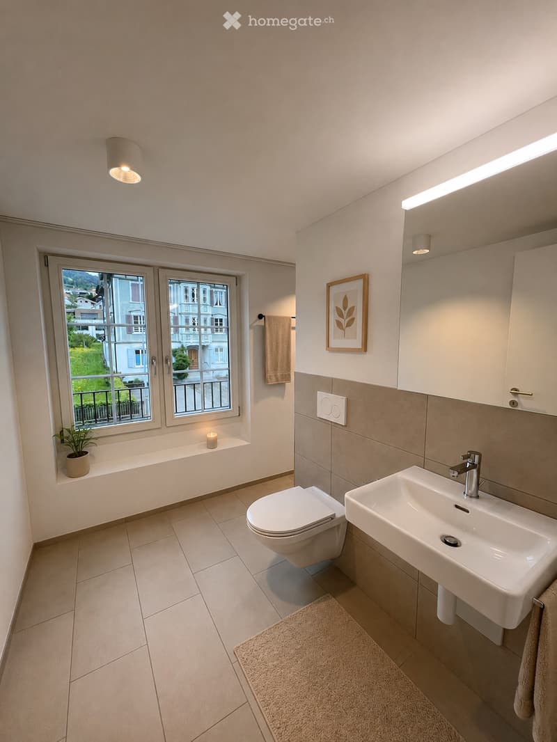 Modern bathroom with a toilet, sink, and a window offering a view of the outside. Includes a towel, a small plant, a picture frame, and a rug on the floor.