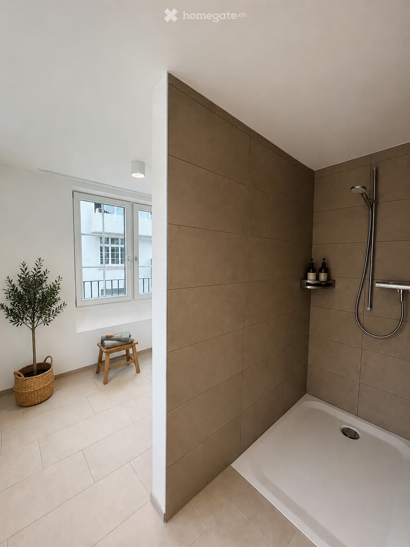 Modern bathroom with a bathtub, shower, and a small wooden stool with towels. A potted plant is placed near the window, and the space features light-colored tiles and a sleek design.