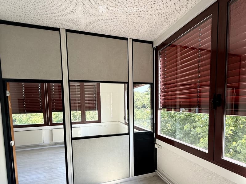 Empty room with large windows, wooden blinds, and a view of greenery outside. The room has a tiled floor and white walls.