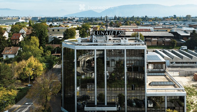 A modern, multi-story building with reflective glass windows and a rooftop sign reading "LUNAJETS." The structure is surrounded by trees, other buildings, and a parking area with several cars.