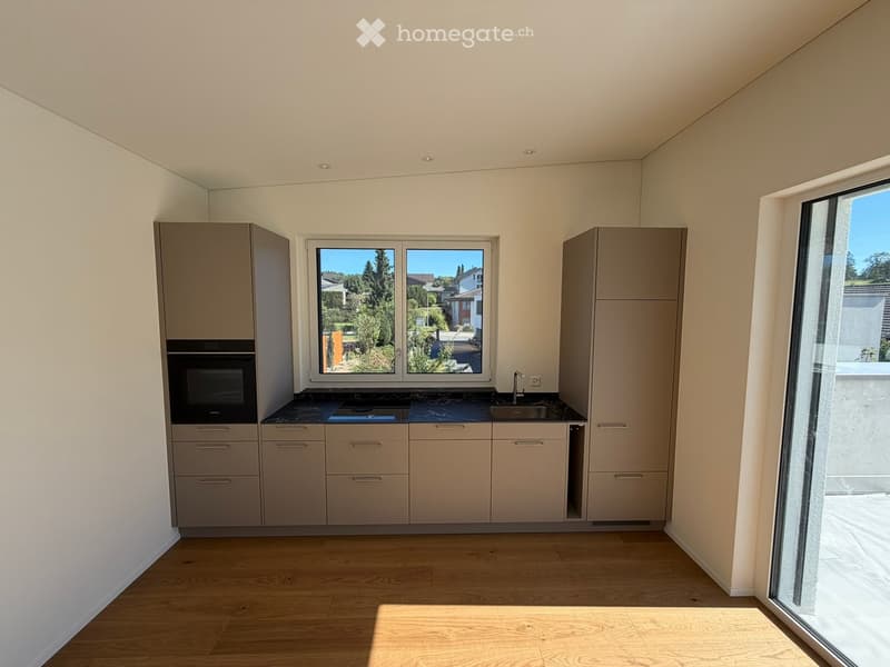 Modern kitchen with light-colored cabinets, a central island, and a large window overlooking a garden. The floor is wooden, and the countertops are dark.