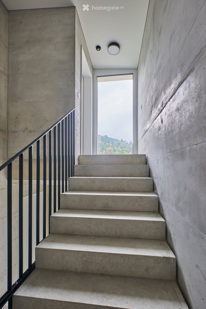 Concrete staircase with modern black metal railing, natural light from a window at the top, and recessed lighting fixtures on the ceiling.