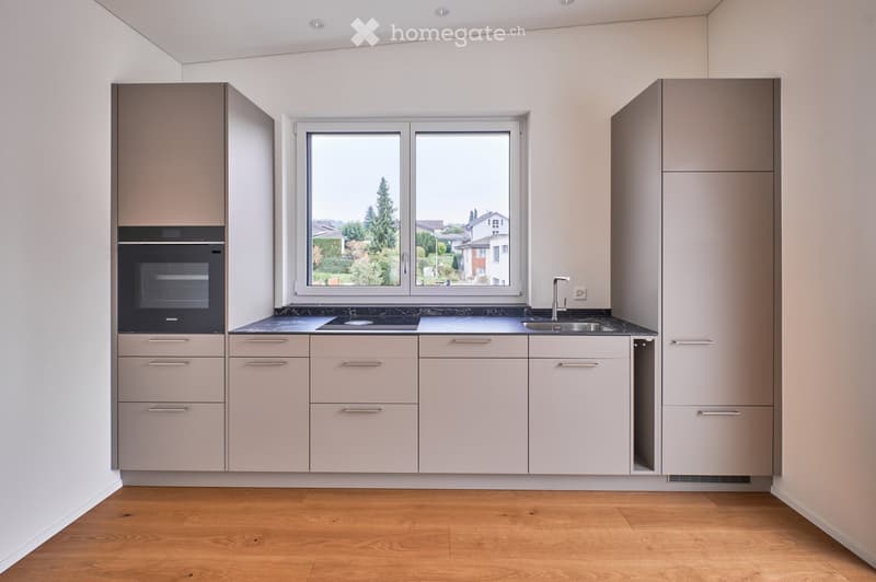 Modern kitchen with light grey cabinets, a central island, and a large window overlooking a green area.