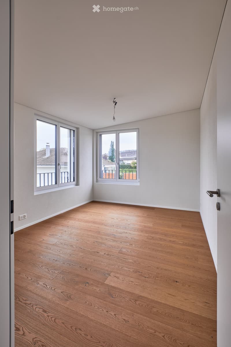 Empty room with light wooden flooring, white walls, and large windows offering a view of the outside. The room has a modern design with a visible light fixture and a door handle.