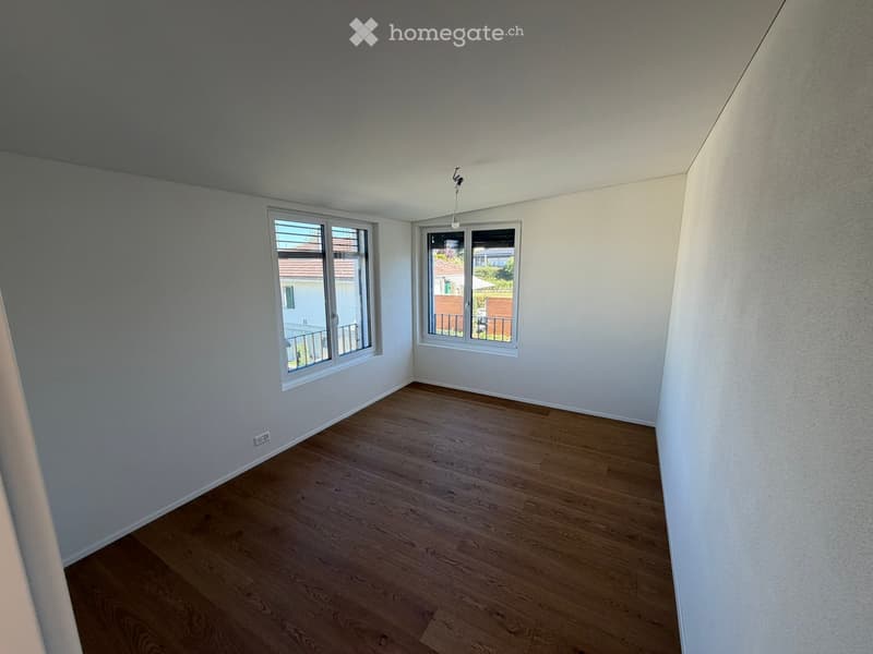 Empty room with wooden flooring, white walls, and two windows offering a view of neighboring houses and a balcony.