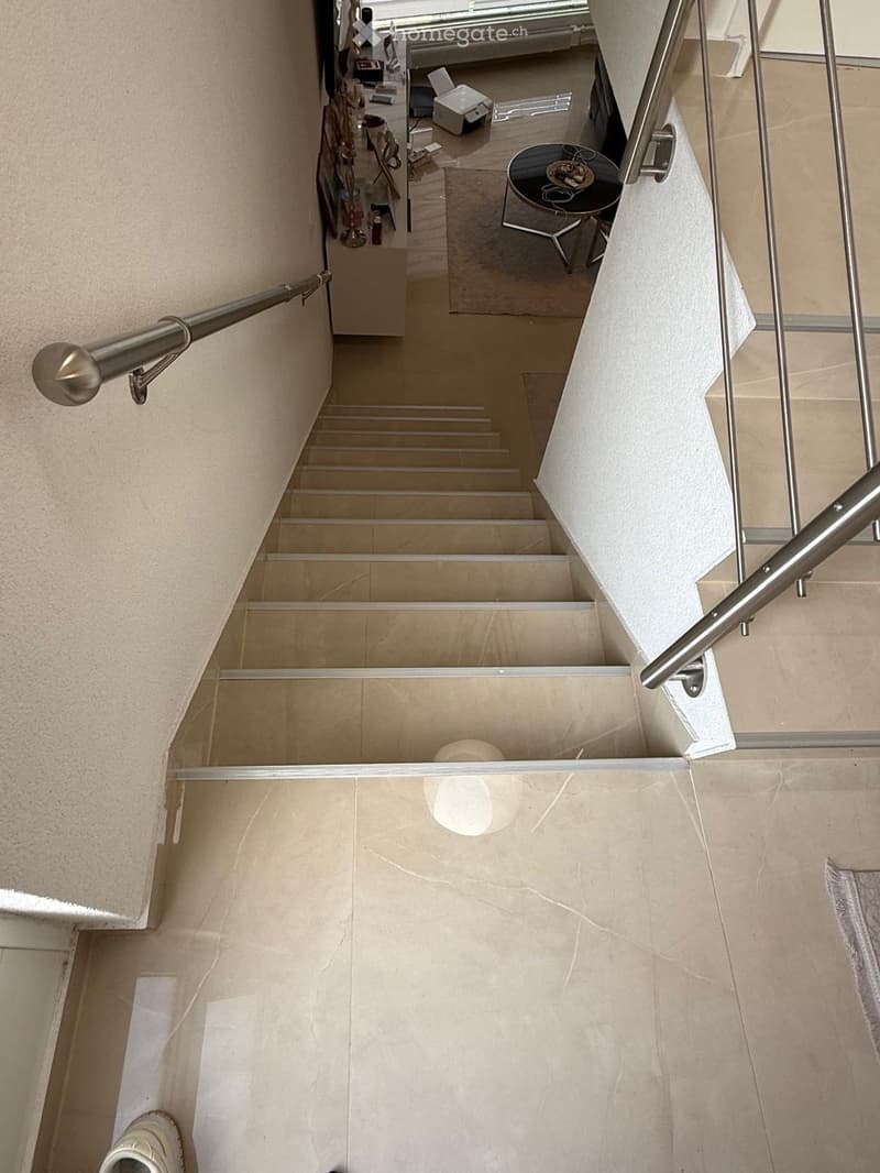 Modern staircase with beige marble steps and polished metal railings, leading to an open area with a small round table and chairs visible in the background.