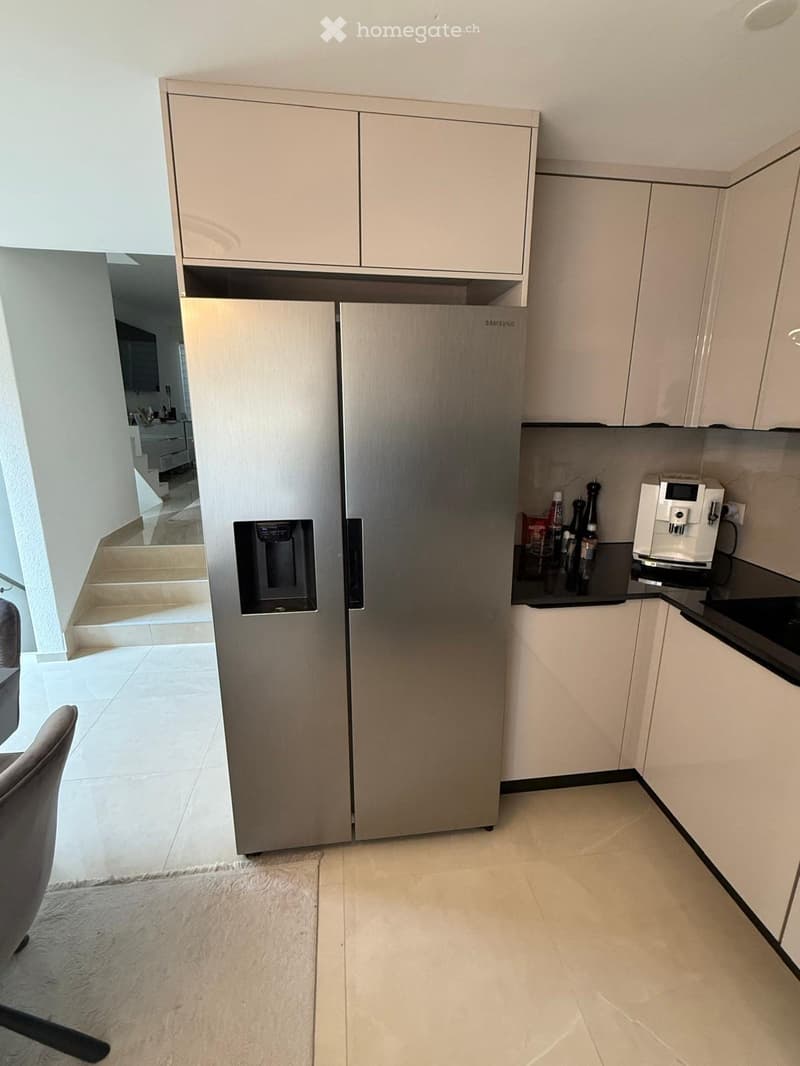Modern kitchen with light-colored cabinets, stainless steel refrigerator, coffee maker on counter, and a small rug on the floor.