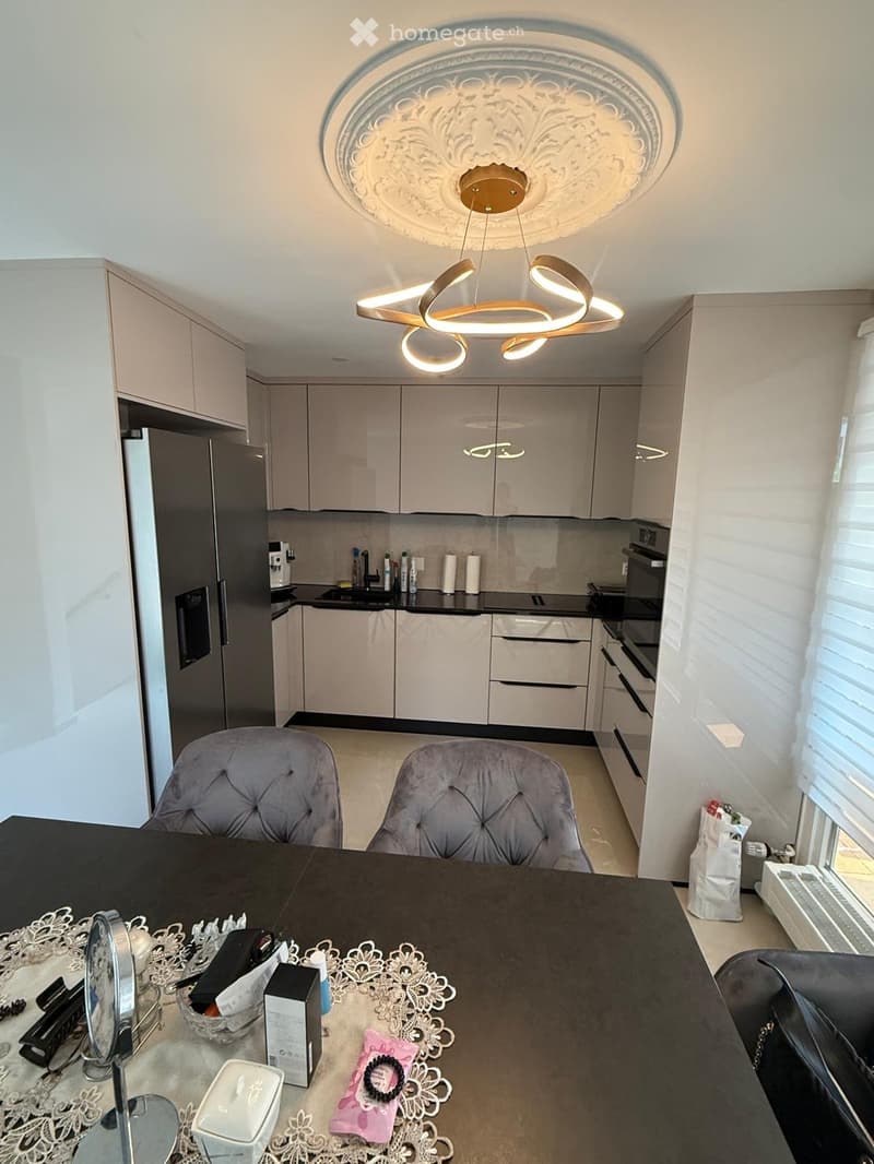 Modern kitchen with light grey cabinets, black countertops, and a sleek chandelier. There is a dining table with two chairs, a refrigerator, and various kitchen appliances.