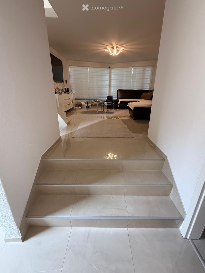 A modern hallway with tiled floors, a staircase leading upwards, and a chandelier on the ceiling. The hallway is well-lit with natural light coming from a large window at the end.