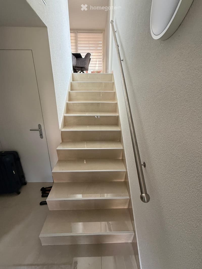 Staircase with beige stone steps, metal handrail, and a view of a room with a chair and desk in the background.