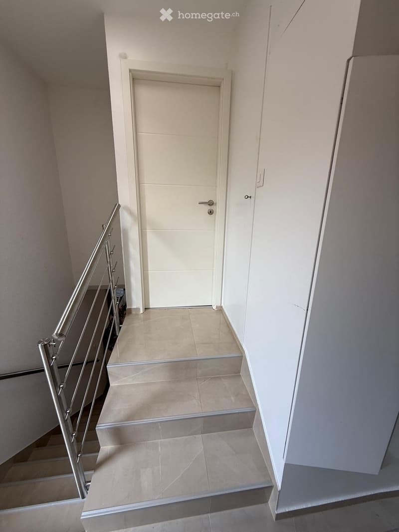 A modern staircase with beige tiled steps and a sleek metal railing, leading up to a closed white door.