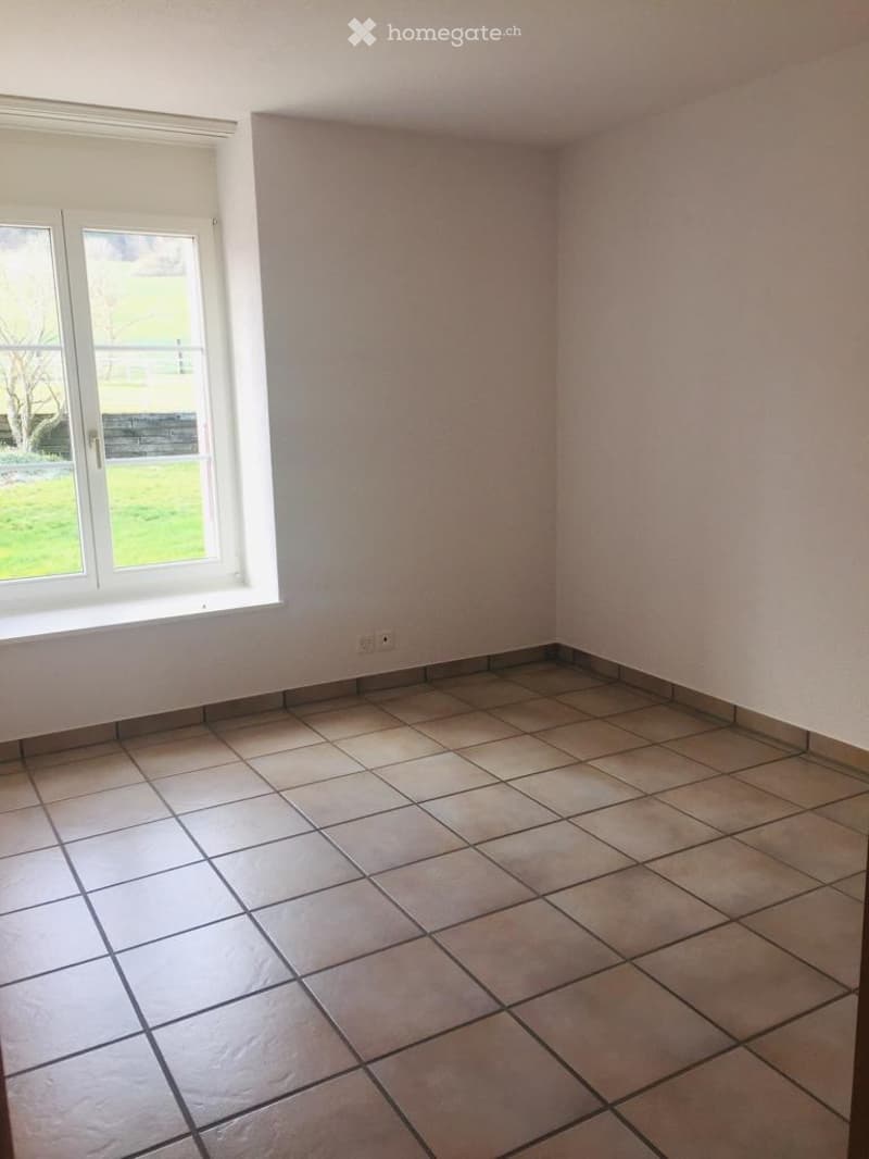 Empty room with tiled floor and white walls, featuring a large window with white frame and a view of a green outdoor area.