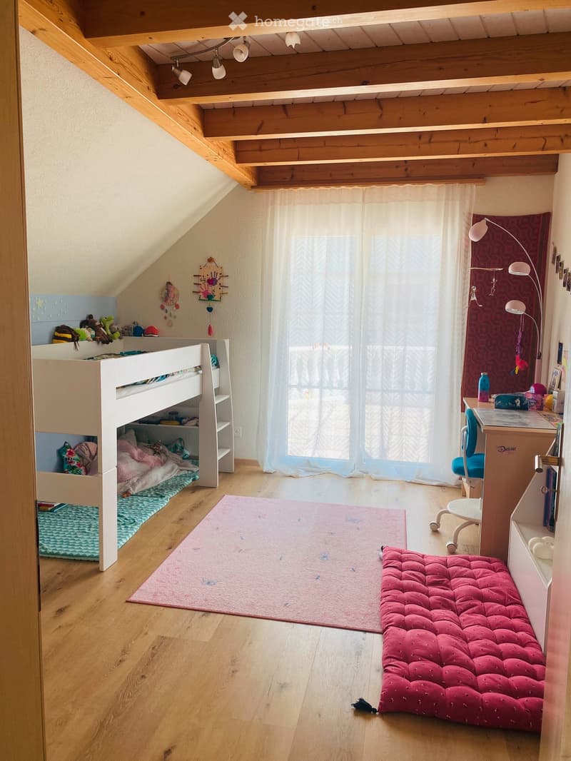 Cozy attic bedroom with wooden slanted ceilings, bunk bed, desk, and a red tufted bench. Features a large window with sheer curtains and a colorful rug.