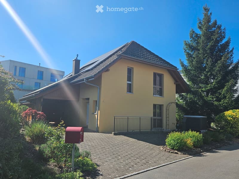 Single-story house with a yellow exterior, dark roof, and a small garage. The house has multiple windows, a chimney, and is surrounded by greenery and a well-maintained garden.