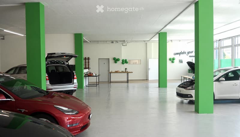 Modern showroom with several cars displayed, including a red Tesla and a white Volkswagen. Green pillars, white walls, and large windows provide natural light. Small tables and shelves are present, along with decorative green spherical objects on the wall