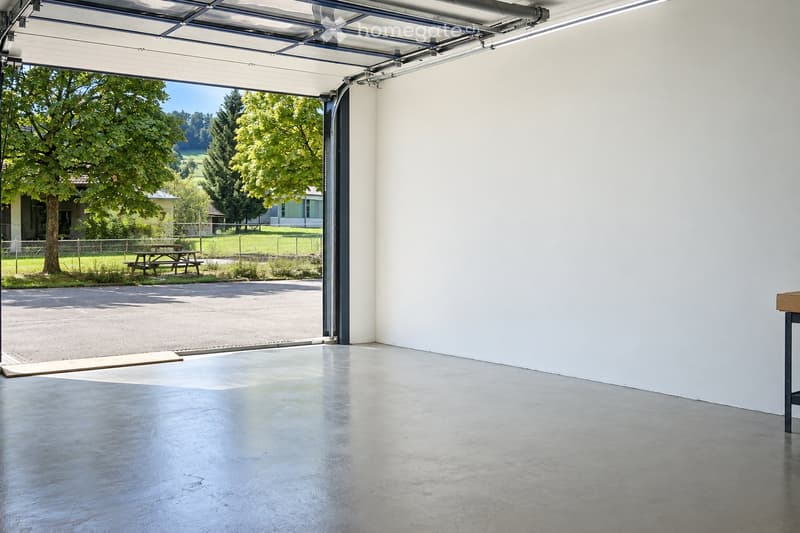Empty garage with a large, open garage door leading to a grassy area with trees and a picnic table outside.