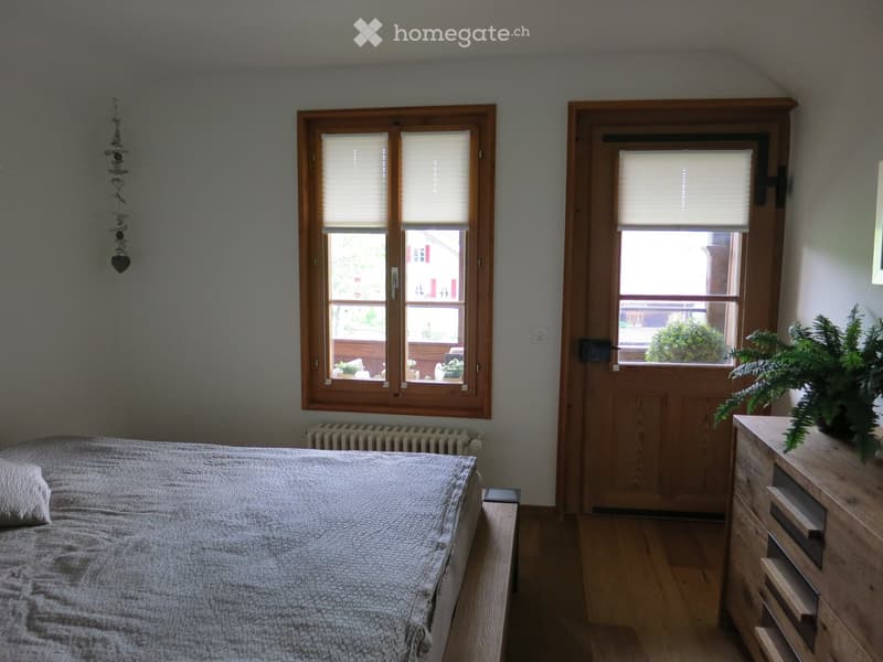 Cozy bedroom with a wooden bed, nightstand, dresser, and plants near the window. The room features wooden flooring, a radiator, and a door leading outside.