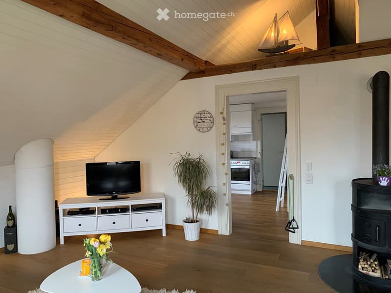 A cozy living room with wooden flooring, a television on a white stand, a coffee table with flowers, and a view into a kitchen area with white cabinets and an oven.