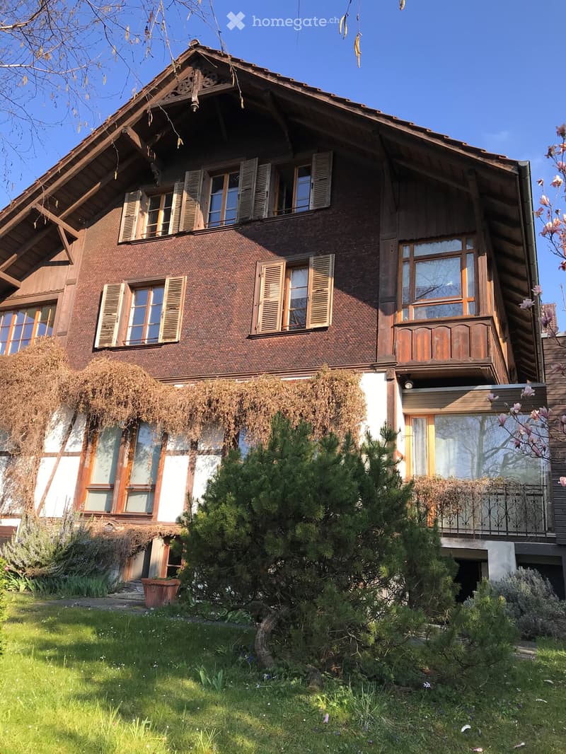 Two-story wooden house with a steeply pitched roof, multiple windows with shutters, and a balcony on the upper floor. The house is surrounded by greenery, including trees and shrubs, and has a well-maintained lawn in front.