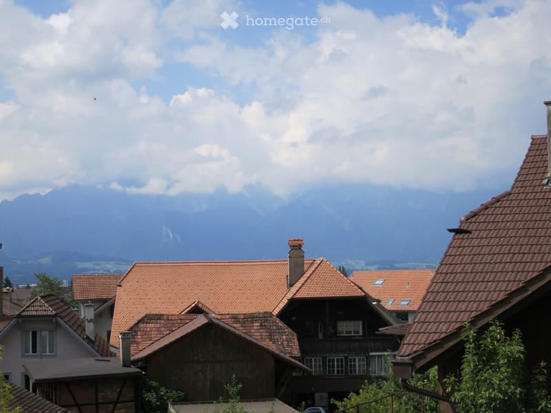 A cluster of traditional Swiss houses with red-tiled roofs and wooden facades, set against a backdrop of mountains and a partly cloudy sky.
