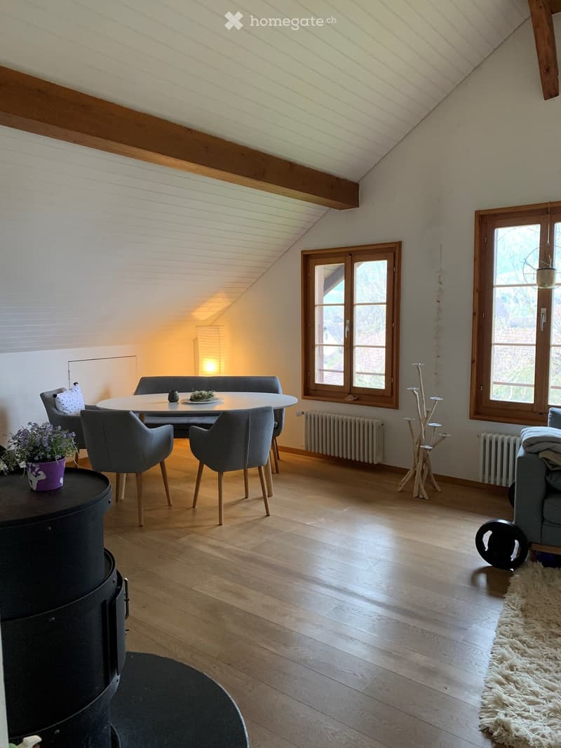 Bright, wooden-floored room with slanted ceiling, large windows, sofa, dining table with chairs, flower pot, lamp, radiator, and decorative stool.