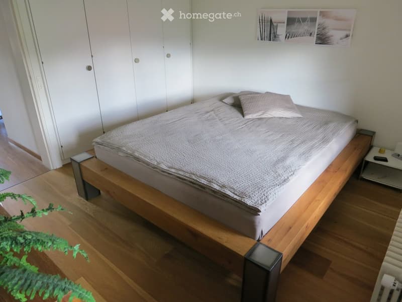 Modern bedroom with a wooden bed frame, grey bedding, nightstand, and a mirror on the wall.