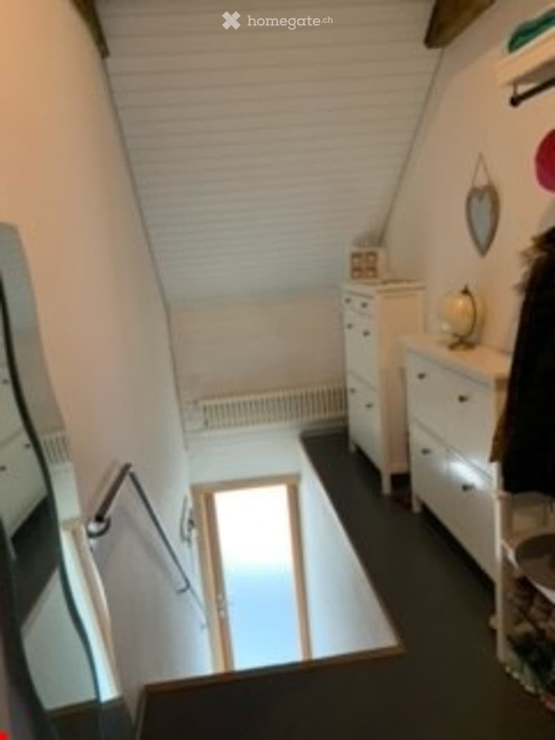 Small, slanted-ceiling room with a single chair, a dresser, a mirror, a radiator, and a shelf with items.