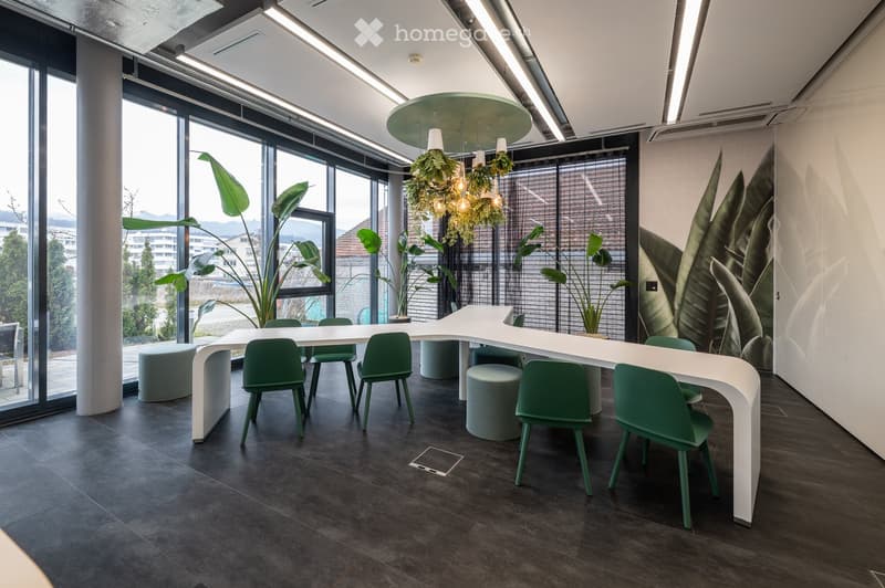 Modern office space with a white curved table, green chairs, grey stools, large windows, hanging plants, and a leafy mural on the wall.