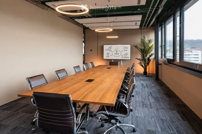 Modern conference room with a long wooden table surrounded by black leather chairs, a large screen on the wall, and a potted plant near the window.