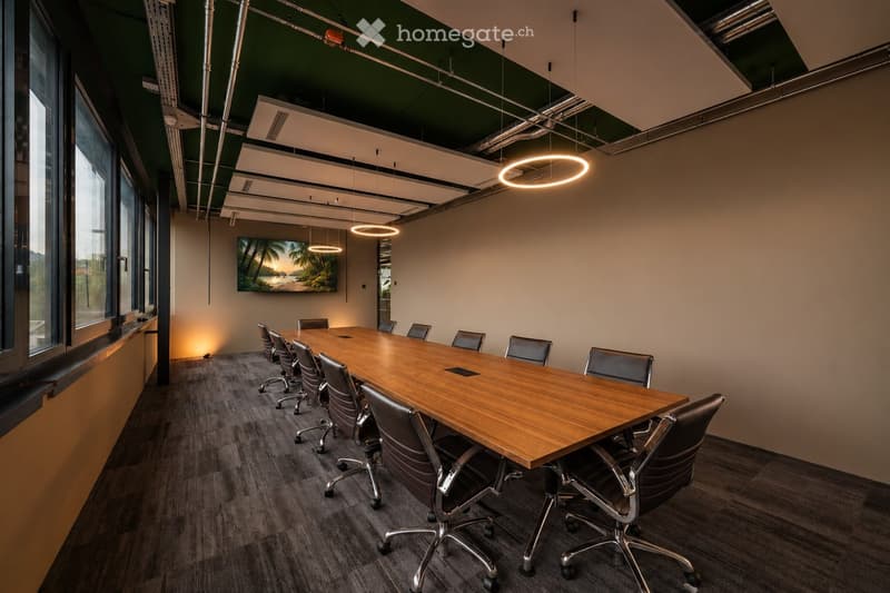 Modern conference room with a long wooden table, multiple chairs, and a large screen on the wall. The room features large windows, pendant lights, and a neutral color palette.
