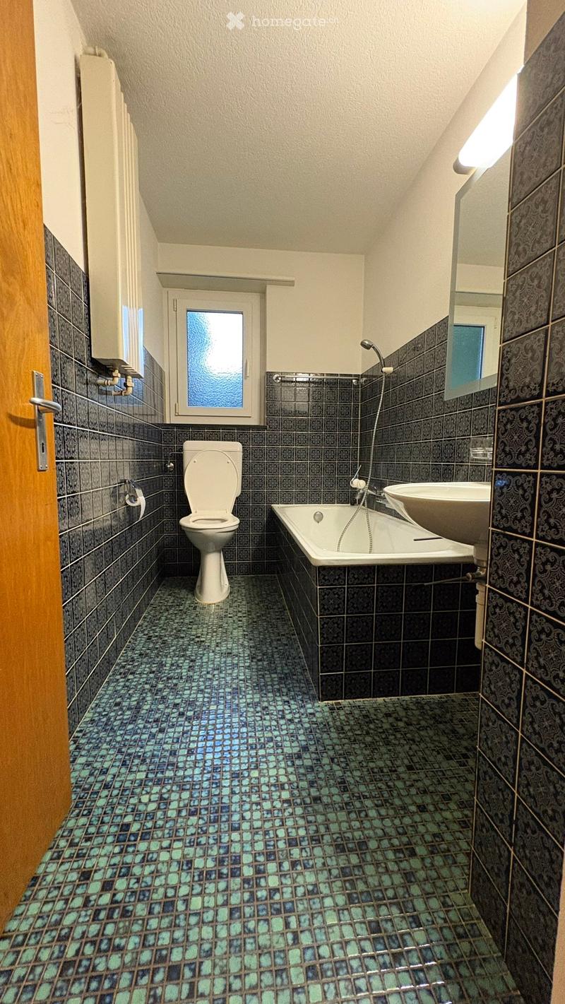Bathroom with dark grey tiles, a toilet, a bathtub, a sink, and a showerhead. The floor features a mosaic of blue and green tiles.