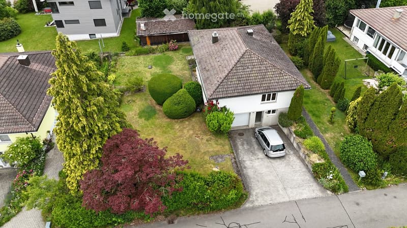 Aerial view of a residential area with multiple houses, well-maintained lawns, trees, and a driveway with a parked car.