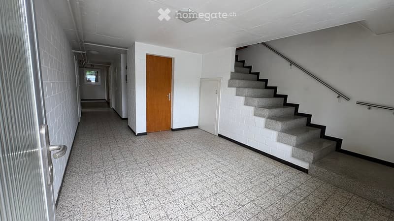 Empty hallway with tiled floor, white walls, a staircase with gray steps, and a wooden door.