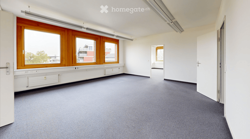 Empty room with grey carpet, white walls, large windows with wooden frames, and a door leading to another space.