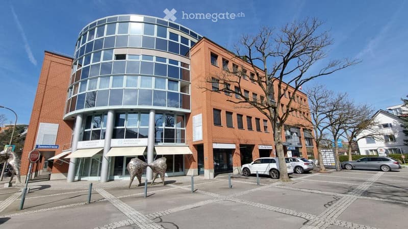 Modern multi-story building with a mix of red brick and glass facade, featuring a rounded glass tower section and several parked cars in front.