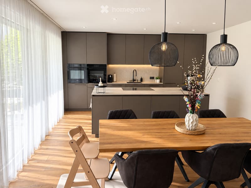 Modern kitchen with grey cabinets, a central island, and a wooden dining table surrounded by chairs. Features pendant lights, a sink, and decorative vase on the table.