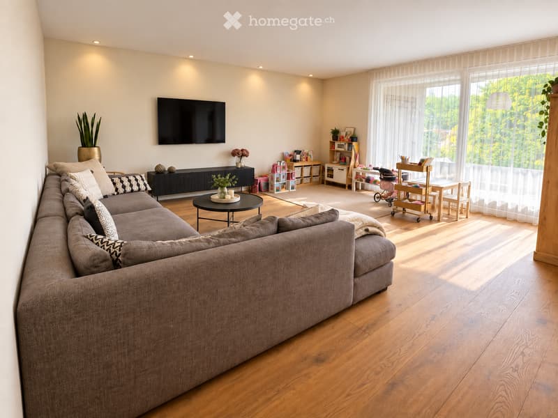 Modern living room with a large sectional sofa, coffee table, TV mounted on the wall, sideboard, dining table with chairs, baby stroller, and storage unit. The room features wooden flooring and large windows with sheer curtains.